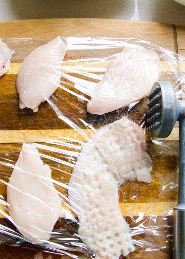 Chicken breasts covered with plastic wrap with meat mallet on cutting board.